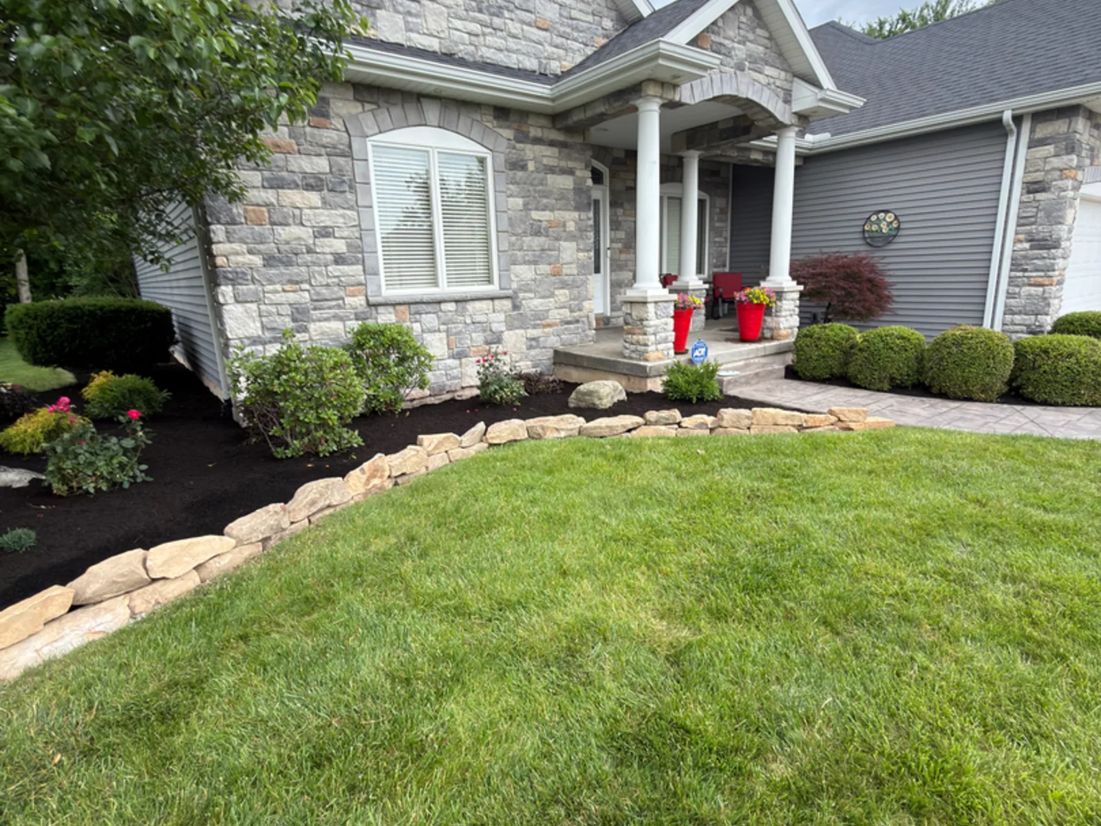 Stone edged landscape bed with fresh mulch and red flowers in front of a stone faced home